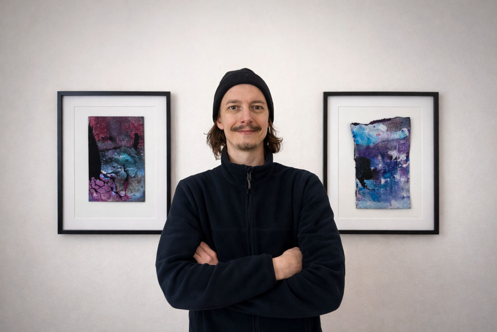 Chris Cabrim in seinem Atelier in Köln-Ehrenfeld vor zwei gerahmten Arbeiten aus der Werkgruppe Other Worlds.